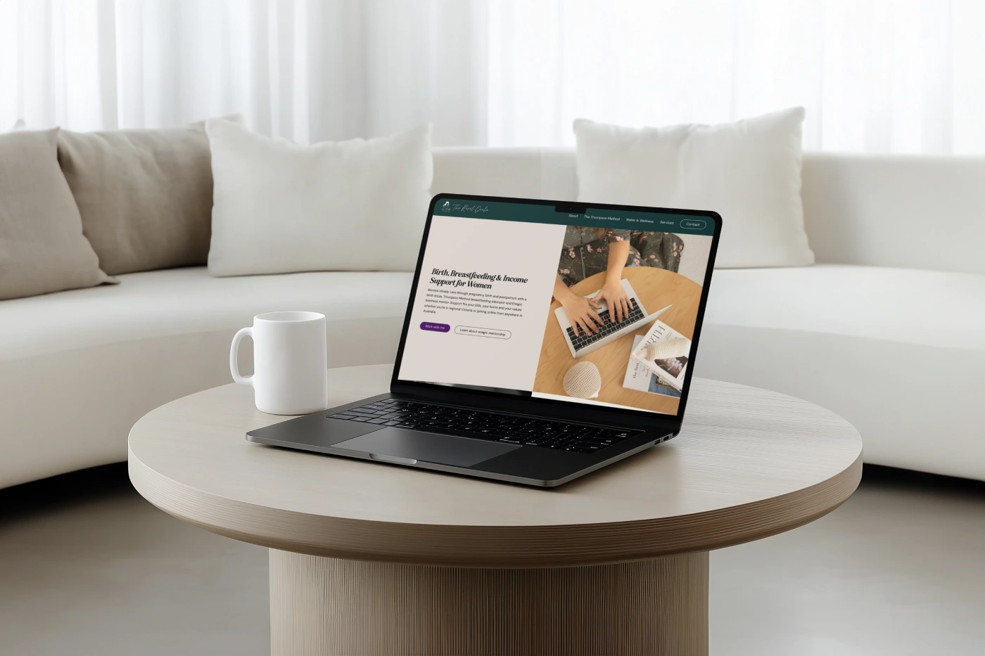 Laptop displaying "The Rural Doula" website on a coffee table, with a mug and cozy seating in the background.